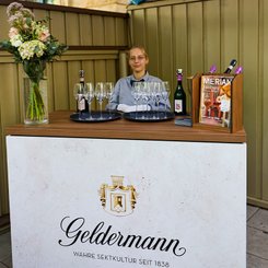 Eine junge Dame bietet die Bayreuther Festspiel Cuvée 2025 am Geldermann Sektstand während der Bayreuther Festspiele an