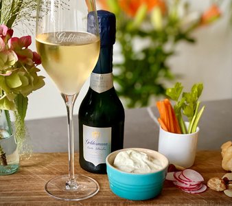 Ein elegantes Glas Sekt steht neben einer Flasche Geldermann, umgeben von frischem Gemüse und einer cremigen Dip-Schale.