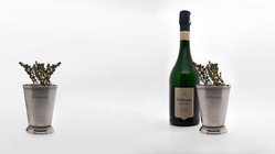 Auf einem minimalistischen Hintergrund sind ein silberner Becher mit Kräutern und eine Flasche Geldermann Sekt zu sehen.