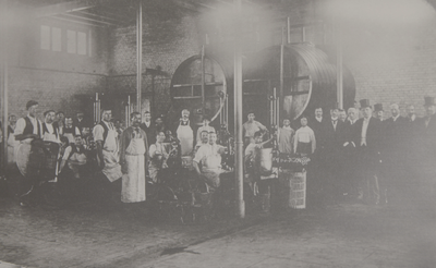 Männer und Frauen in Arbeitskleidung posieren in einer industriellen Umgebung, umgeben von großen, runden Maschinen.