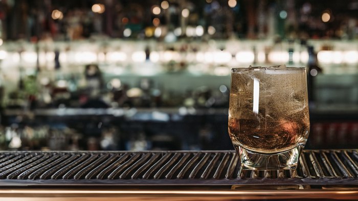 Das Glas mit Eis und einem erfrischenden Getränk reflektiert das Licht, während die Bar im Hintergrund eine lebhafte Atmosphäre schafft.