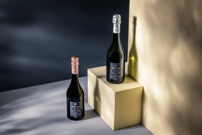 Zwei Flaschen Crémant stehen auf einem hellen Würfel, beleuchtet von sanften Schatten und einem dunklen Hintergrund.