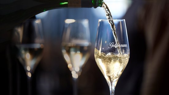 Ein Glas wird mit sprudelndem Sekt gefüllt, während im Hintergrund verschwommene Gläser zu sehen sind.