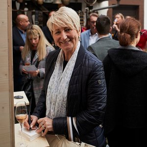Eine lächelnde Frau mit blonden Haaren hält einen Stift und notiert etwas auf einem Block, während sie in einer Menschenmenge steht.