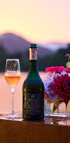 Geldermann Soirée Rosé: Eine Flasche Geldermann Crémant Baden und ein Glas Rosé Sekt vor der Kulisse des Tegernsees im Abendrot