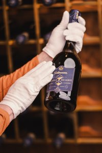Eine Person in weißen Handschuhen hält eine Flasche Sekt mit einem eleganten Etikett in einem Weinkeller.