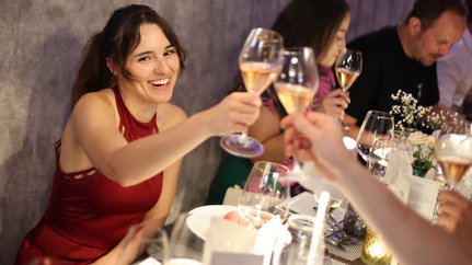 Eine fröhliche Frau in einem roten Kleid hebt ihr Glas in einem festlichen Ambiente, um auf einen besonderen Moment anzustoßen.
