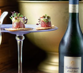 Ein stilvolles Arrangement mit zwei kleinen Häppchen auf einem Glasuntersetzer, begleitet von einer Flasche Geldermann Sekt.