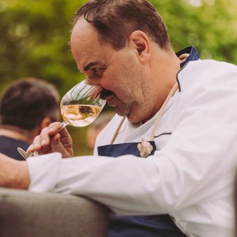 Ein Mann mit Schnurrbart und Kochjacke riecht an einem Glas Wein, während eine Frau mit langen Haaren und rotem Lippenstift lächelt.