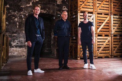 Die drei Männer, gekleidet in lässiger Kleidung, stehen in einem alten Lagerraum und strahlen Freude und Kameradschaft aus.