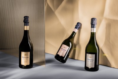 Drei Flaschen Geldermann-Sekt stehen auf einem eleganten Tisch, umgeben von sanften Lichtreflexionen und einem strukturierten Hintergrund.