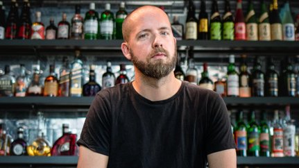 Der Bartträger in einem schwarzen T-Shirt wirkt nachdenklich, während er auf einer Bar sitzt, die mit Flaschen dekoriert ist.