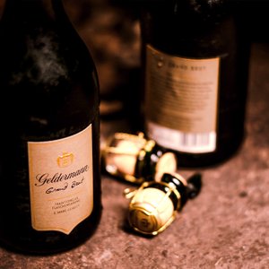 Eine Flasche Geldermann Grand Brut steht im Vordergrund, während zwei Korken mit goldenen Kappen im Hintergrund liegen.