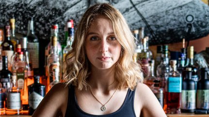 Eine junge Frau mit blonden Locken steht hinter einer Bar, umgeben von einer Vielzahl von Spirituosen und Gläsern.