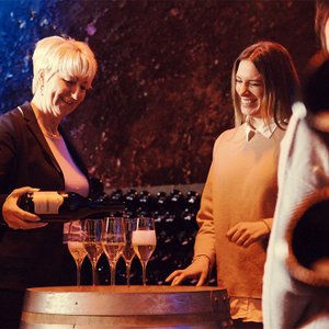 Eine blonde Frau schenkt Sekt in Gläser, während eine andere Frau lächelt und die Szene genießt, umgeben von Weinfässern.