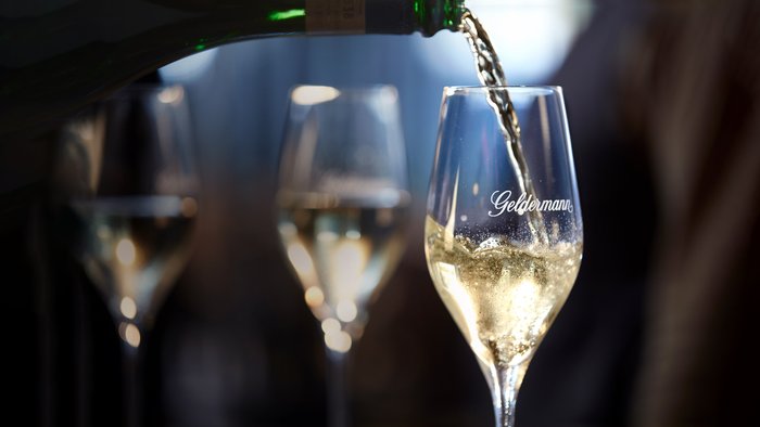 Ein Glas wird mit sprudelndem Sekt gefüllt, während im Hintergrund verschwommene Gläser zu sehen sind.