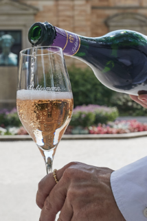 Ein Glas Sekt wird von einer Hand aus einer Flasche mit lila Etikett eingeschenkt, umgeben von blühenden Gärten.