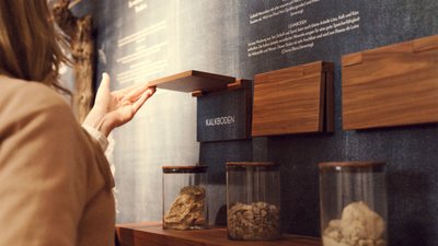 Eine Person hebt eine Holzplatte an, um Informationen über verschiedene Bodenarten zu entdecken, während Gläser mit Proben daneben stehen.