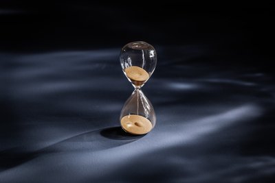 Eine elegante Sanduhr steht auf einer dunklen Oberfläche, das Licht reflektiert sanft auf dem Glas und dem Sand.