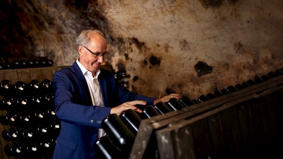 Ein Mann in einem Anzug lächelt, während er sorgfältig an einer Reihe von Weinflaschen in einem Keller arbeitet.