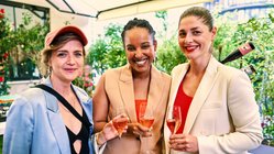 Mit Gläsern in der Hand posieren drei Frauen in modischen Outfits vor einer blühenden Kulisse und genießen den Moment.