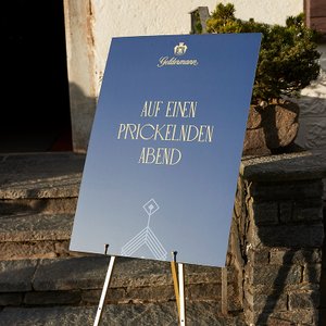 Das dekorative Schild mit dem Motto 'Auf einen prickelnden Abend' steht auf einem stabilen Ständer vor einem Eingang.