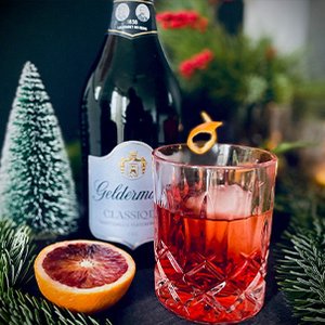 Ein elegantes Glas mit einem roten Getränk, garniert mit einer Orangenscheibe, steht neben einer Flasche Sekt.