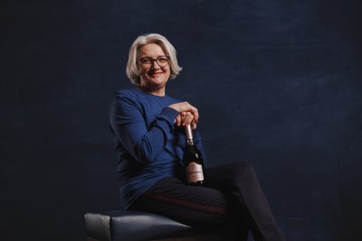 Eine lächelnde Frau mit grauen Haaren sitzt auf einem Hocker und hält eine Flasche Sekt in der Hand.