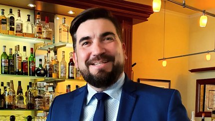 Ein lächelnder Mann mit Bart trägt einen Anzug und steht vor einer Bar mit einer Vielzahl von Spirituosen im Hintergrund.
