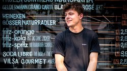 Der Mann trägt ein schwarzes T-Shirt und kurze Hosen, während er lächelnd auf einer Erhöhung sitzt, die mit Graffiti verziert ist.