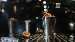 Ein eleganter silberner Becher wird mit einem sprudelnden Getränk und einer Scheibe Orange gefüllt, während Eiswürfel sichtbar sind.