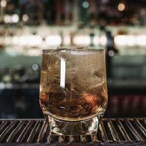 Ein Glas mit einem sprudelnden Getränk, gefüllt mit Eiswürfeln, steht auf einer Bar mit verschwommenem Hintergrund.