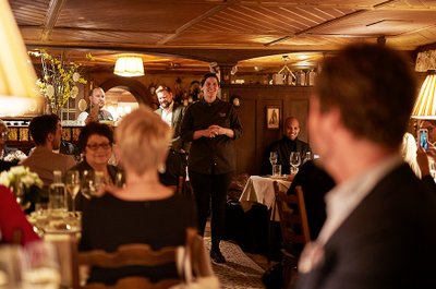 Eine lächelnde Köchin spricht zu den Gästen in einem gemütlichen Restaurant mit warmem Licht und rustikalem Ambiente.