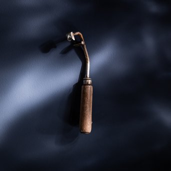 Ein gebrauchtes Werkzeug mit einem Holzgriff und einem metallischen Kopf hängt vor einem dunklen, strukturierten Hintergrund.