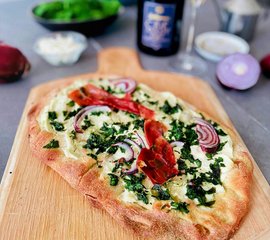 Eine frisch gebackene Pizza auf einem Holzbrett, belegt mit roten Zwiebeln, Schinken und frischem Blattgrün.