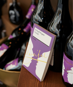 Im Vordergrund ist ein Buch mit dem Titel 'Parsifal' zu sehen, umgeben von glänzenden Weinflaschen auf einem Holzregal.