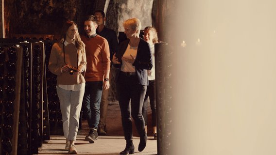 Im Weinkeller unterhalten sich vier Personen lebhaft, während eine Frau mit einer Kamera in der Hand lächelt.