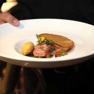Ein feines Gericht, das rosa Fleisch, eine gelbe Kugel und knuspriges Brot zeigt, präsentiert in einem modernen, eleganten Stil.