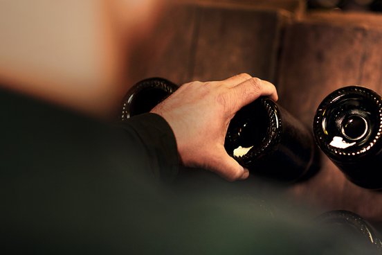 Eine Person berührt eine Flasche, die auf einer Holzoberfläche steht, umgeben von weiteren Flaschen im Hintergrund.