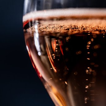 Ein Glas mit sprudelndem, roséfarbenem Getränk, das in einem dunklen Hintergrund schimmert und Blasen aufsteigt.