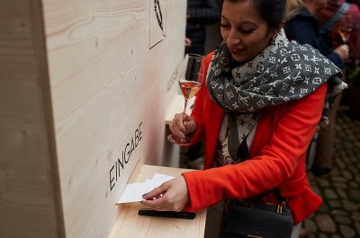Eine Frau in einem leuchtend roten Mantel hält ein Glas Sekt und legt eine Karte auf einen hölzernen Tisch.