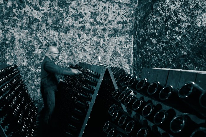In einem dunklen Weinkeller arbeitet ein älterer Mann an der Anordnung von Flaschen, während eine Kerze schwach leuchtet.