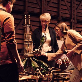 In einem rustikalen Raum arbeiten drei Personen an einer Apparatur, die Flaschen mit einem mechanischen System befüllt.