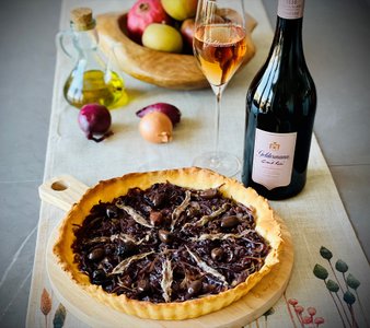 Der Zwiebelkuchen, reich belegt mit schwarzen Oliven und Sardellen, steht auf einem Tisch mit frischen Äpfeln und Zwiebeln.