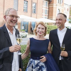 Ein älterer Mann, eine Frau mit roten Haaren und ein jüngerer Mann lächeln fröhlich, während sie an einem festlichen Anlass teilnehmen.