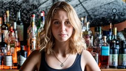 Eine junge Frau mit blonden Locken steht hinter einer Bar, umgeben von einer Vielzahl von Spirituosen und Gläsern.