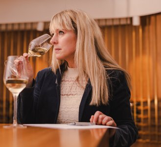 Eine Frau mit langen blonden Haaren hält ein Weinglas und betrachtet es nachdenklich, während sie an einem Tisch sitzt.