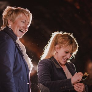 Zwei Frauen lachen fröhlich, während eine von ihnen eine Sektflasche öffnet, umgeben von einer festlichen Atmosphäre.