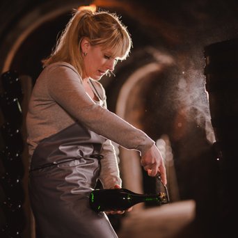 In einem dunklen Weinkeller steht eine blonde Frau mit einem Korkenzieher, bereit, eine Flasche Sekt zu öffnen, umgeben von Weinflaschen.