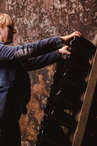 Mit einem konzentrierten Ausdruck stellt eine Person Flaschen auf ein Regal, umgeben von einer rustikalen, abgenutzten Wandstruktur.